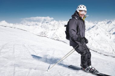 Bir kask ve kar maskesi ile sakallı erkek kayakçı kar kaplı dağların arka plan ve mavi bir gökyüzüne karşı kayaklar üzerinde kar tozu ile frenler. Siyah takım elbiseli atlet