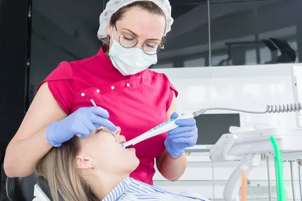 Uma dentista profissional de óculos e macacão examina a cavidade oral de uma jovem na cadeira dental usando uma câmera de vídeo estomatológica intraoral com iluminação LED — Fotografia de Stock