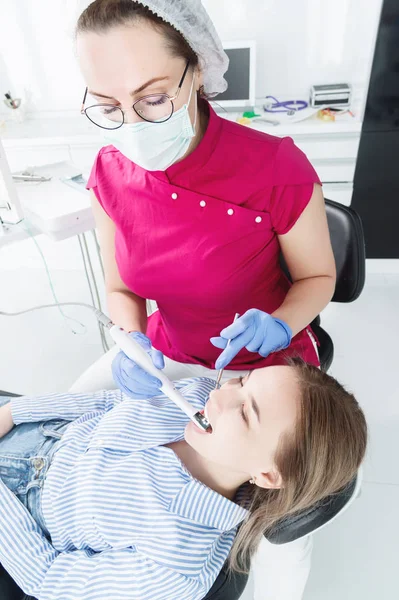 Μια επαγγελματική γυναίκα οδοντίατρος σε γυαλιά και φόρμες εξετάζει την στοματική κοιλότητα ενός νεαρού κοριτσιού στην πολυθρόνα με χρήση ενδοστοματικής σταμολογικής βιντεοκάμερας με φωτισμό LED — Φωτογραφία Αρχείου