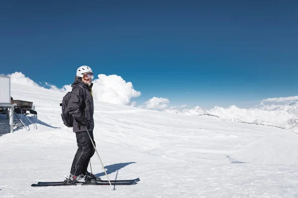 Um esquiador barbudo em um capacete e uma máscara de esqui está de pé em esquis contra o fundo de montanhas cobertas de neve e um céu azul. Atleta de terno preto — Fotografia de Stock
