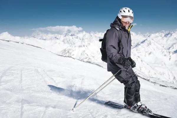 Brodaty mężczyzna narciarz w kasku i maski narciarskie hamulce z śniegu w proszku na nartach na tle ośnieżonych gór i błękitne niebo. Sportowiec w czarnym garniturze — Zdjęcie stockowe