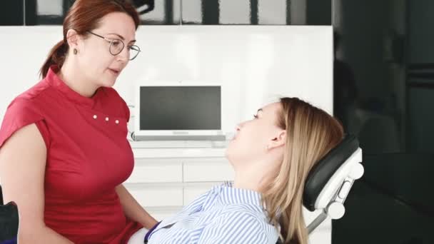 Una joven en una consulta con una mujer dentista en gafas está sentada en una silla en una oficina de estamotología. La conversación del médico y el paciente — Vídeos de Stock