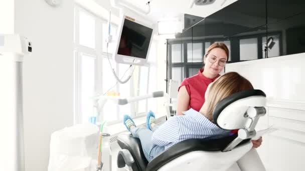 Una joven en una consulta con una mujer dentista en gafas está sentada en una silla en una oficina de estamotología. La conversación del médico y el paciente — Vídeo de stock