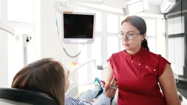 Una joven en una consulta con una mujer dentista en gafas está sentada en una silla en una oficina de estamotología. La conversación del médico y el paciente — Vídeo de stock
