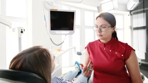 Una joven en una consulta con una mujer dentista en gafas está sentada en una silla en una oficina de estamotología. La conversación del médico y el paciente — Vídeos de Stock