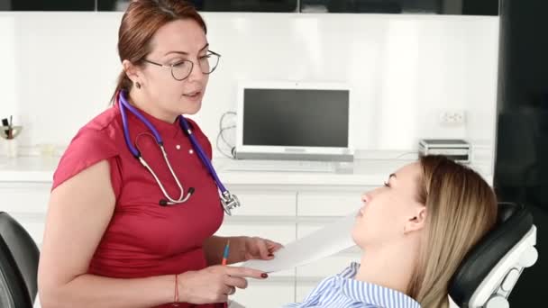 Una joven en una consulta con una mujer dentista en gafas está sentada en una silla en una oficina de estamotología. La conversación del médico y el paciente — Vídeo de stock