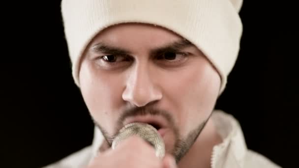 Κοντινό-up τραγουδιστή ροκ ποπ με ένα κομψό μούσι σε λευκά ρούχα και ένα καπέλο με ένα μικρόφωνο στα χέρια του έντονα επιθετικά τραγουδώντας στο στούντιο με φόντο τους μαύρους τοίχους — Αρχείο Βίντεο