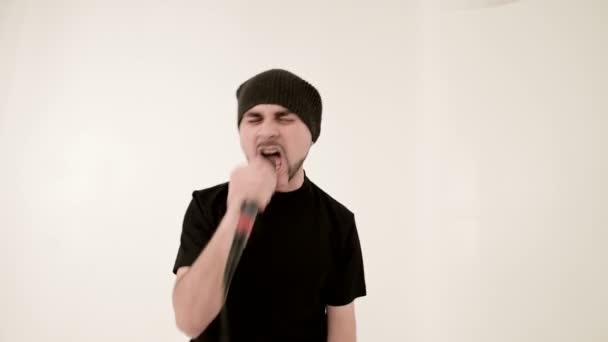 Фронтмен вокаліст рок-поп зі стильною бородою в чорному одязі і капелюхом з мікрофоном в руках виразно агресивно співає в студії на тлі білих стін — стокове відео