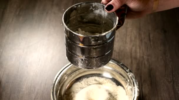 Close-up de uma mão feminina peneirando farinha com uma caneca peneira em uma tigela de metal na cozinha da casa. Cozinhar em casa Cozimento — Vídeo de Stock