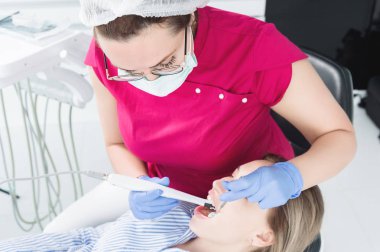 Yakın çekim gözlük ve Tulum bir profesyonel dişçi kadın, LED aydınlatma ile İntraoral stamotological video kamera kullanarak diş sandalyesinde genç bir kızın oral boşluğunu inceliyor