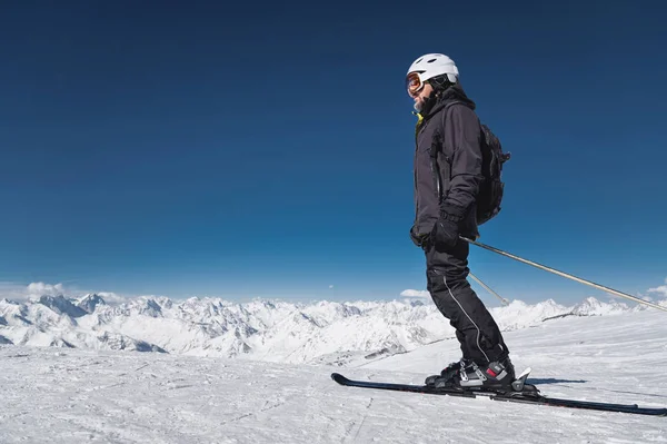 Brodaty mężczyzna narciarz w kasku i maska narciarska stoi na nartach na tle ośnieżonych gór i błękitne niebo. Sportowiec w czarnym garniturze — Zdjęcie stockowe