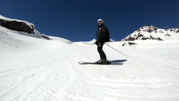 Um esquiador macho de grande ângulo envelhecido em equipamento preto e capacete branco com bastões de esqui cavalga em uma encosta nevada em um dia ensolarado. O conceito de esportes de esqui de inverno — Vídeo de Stock