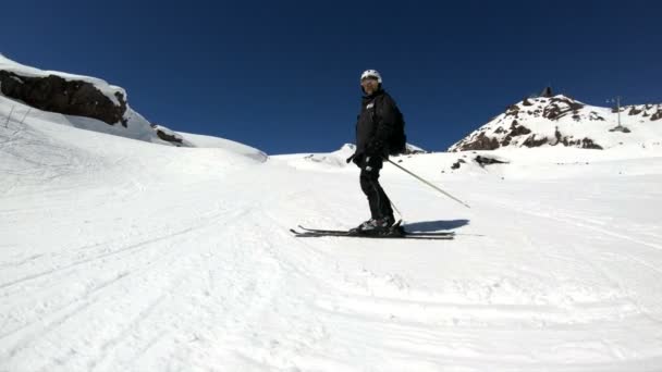 Uno sciatore maschio grandangolare invecchiato in attrezzatura nera e casco bianco con racchette da sci cavalca su un pendio innevato in una giornata di sole. Il concetto di sport sciistici invernali — Video Stock