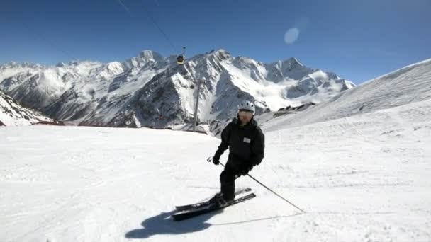 En vidvinkel manliga skidåkare åldrad i svart utrustning och vit hjälm med skidstavar rider på en snöig sluttning på en solig dag. Begreppet vinter skidsport — Stockvideo