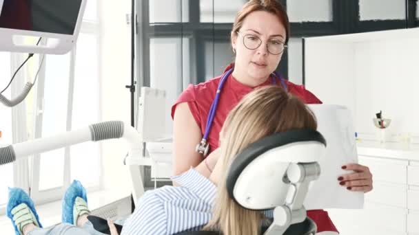 Une jeune fille en consultation avec une dentiste portant des lunettes est assise sur une chaise dans un bureau de stamotologie. La conversation du médecin et du patient — Video