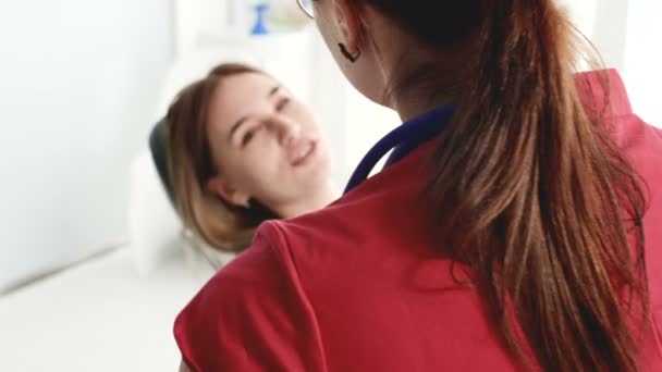 Una joven en una consulta con una mujer dentista en gafas está sentada en una silla en una oficina de estamotología. La conversación del médico y el paciente — Vídeos de Stock