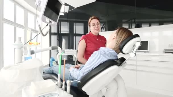En ung flicka vid ett samråd med en tandläkare kvinna i glasögon sitter på en stol i en stamotologi kontor. Samtalet av läkaren och patienten — Stockvideo