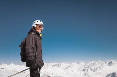 Kasklı ve kar maskeli sakallı bir erkek kayakçı, karla kaplı dağların arka planına ve mavi bir gökyüzüne karşı kayakların üzerinde duruyor. Siyah takım elbiseli atlet