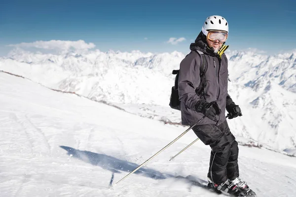 ヘルメットをかぶったひげを生やした男性スキーヤーと、雪に覆われた山々と青い空を背景に、スキーに雪の粉を付けたスキーマスクブレーキ。黒いスーツを着たアスリート — ストック写真