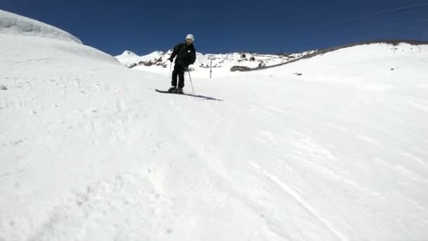 Un skieur masculin grand angle vieilli en équipement noir et casque blanc avec bâtons de ski monte sur une pente enneigée par une journée ensoleillée. Le concept des sports d'hiver — Video