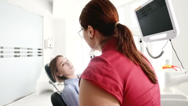 Молодая девушка на консультации с дантисткой в очках сидит на стуле в стоматологическом кабинете. Разговор врача и пациента — стоковое видео