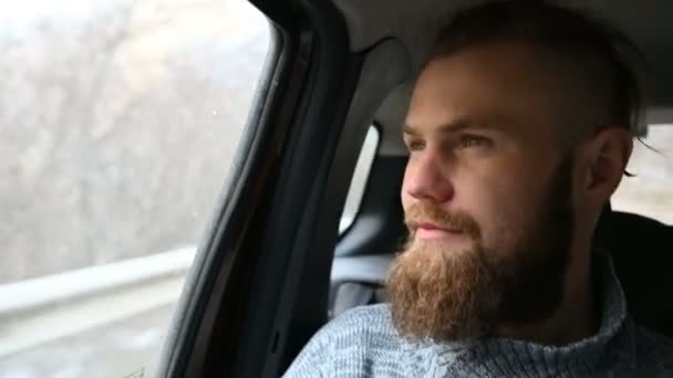 Retrato de cerca de un hombre hipster barbudo riendo sentado en un coche en el asiento trasero y meditando mientras mira a su alrededor — Vídeo de stock