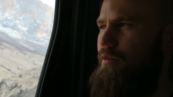 Portrait en gros plan d'un hipster barbu parlant assis dans la voiture sur le siège arrière et méditant sur les côtés sur fond de montagnes — Video