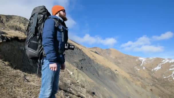 Um homem barbudo de chapéu de óculos de sol com uma mochila e uma câmera fica no alto das montanhas e olha de lado contra o pano de fundo das montanhas cobertas de neve — Vídeo de Stock