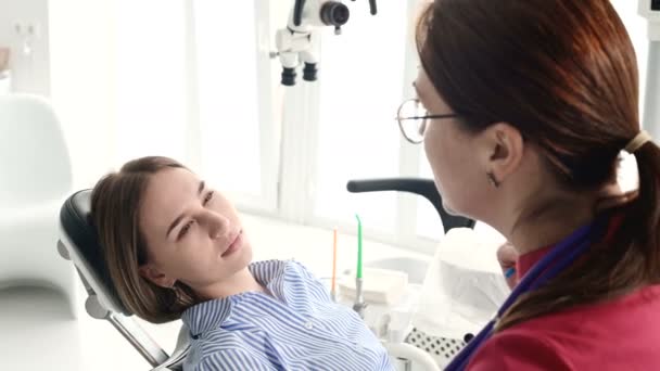 Una joven en una consulta con una mujer dentista en gafas está sentada en una silla en una oficina de estamotología. La conversación del médico y el paciente — Vídeo de stock