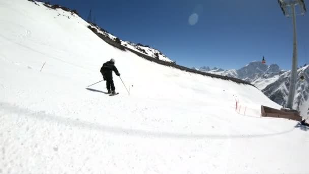 Uno sciatore maschio grandangolare invecchiato in attrezzatura nera e casco bianco con racchette da sci cavalca su un pendio innevato in una giornata di sole. Il concetto di sport sciistici invernali — Video Stock