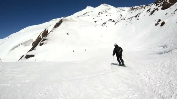 En vidvinkel manliga skidåkare åldrad i svart utrustning och vit hjälm med skidstavar rider på en snöig sluttning på en solig dag. Begreppet vinter skidsport — Stockvideo