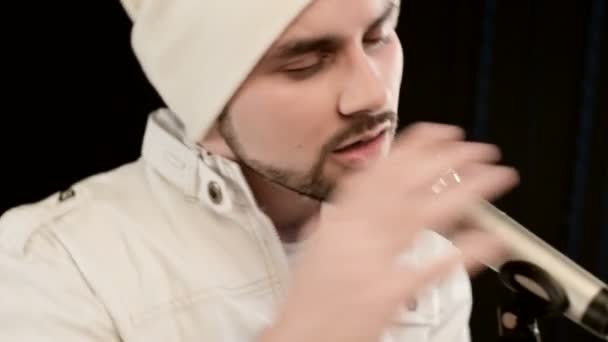 Крупный план Фронтман вокалист рок-поп со стильной бородой в белой одежде и шляпой с микрофоном в руках выразительно агрессивно поет в студии на фоне черных стен — стоковое видео