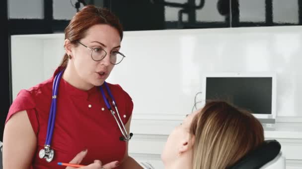 Una joven en una consulta con una mujer dentista en gafas está sentada en una silla en una oficina de estamotología. La conversación del médico y el paciente — Vídeo de stock