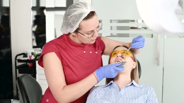 Mooi blond meisje in beschermende gele bril op de stamotoloog onderzocht haar open mond. Vrouwelijke tandarts onderzoekt de mondholte van een jonge patiënt met behulp van een tandarts-instrument — Stockvideo