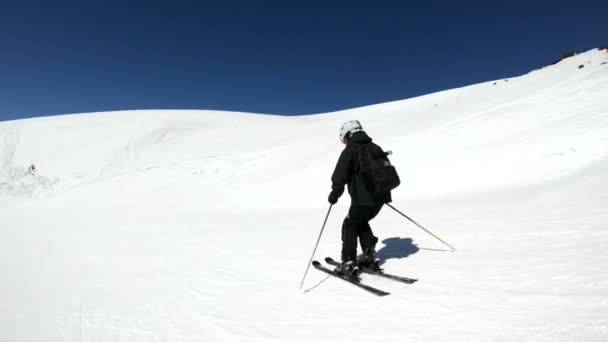 Szeroki kąt męski narciarz w wieku czarnym wyposażeniem i białym hełmem z kijkami jeździ na śnieżnego stoku w słoneczny dzień. Koncepcja zimowych sportów narciarskich — Wideo stockowe