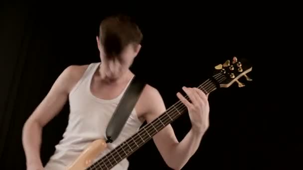 Jovem músico masculino em roupas brancas com um baixo bege em um fundo preto. Baixo guitarrista expressivo jogo de música — Vídeo de Stock