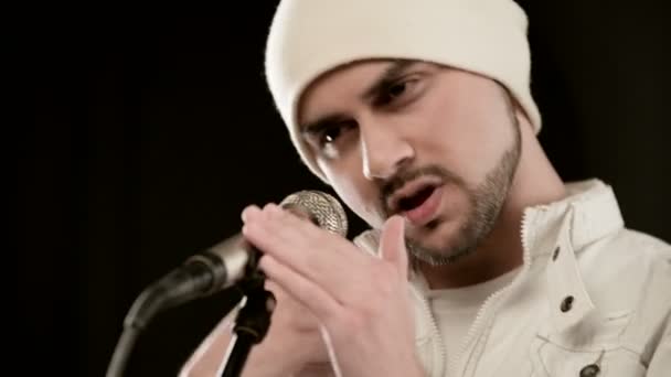 Κοντινό-up τραγουδιστή ροκ ποπ με ένα κομψό μούσι σε λευκά ρούχα και ένα καπέλο με ένα μικρόφωνο στα χέρια του έντονα επιθετικά τραγουδώντας στο στούντιο με φόντο τους μαύρους τοίχους — Αρχείο Βίντεο