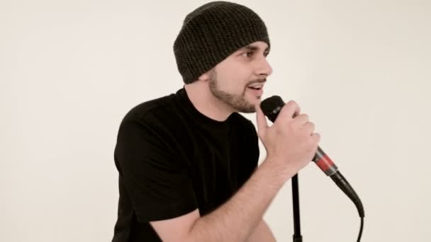 Frontman vocalist rock pop with a stylish beard in black clothes and a hat with a microphone in his hands expressively aggressively singing in the studio against the background of white walls — Stock Video