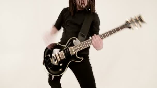 Elegante guitarrista solista con rastas en la cabeza y con ropa negra sobre un fondo blanco tocando expresamente la guitarra negra en un estudio blanco — Vídeos de Stock
