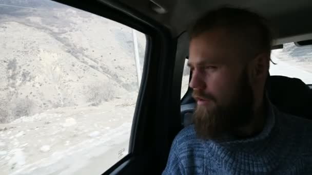 Ritratto ravvicinato di un maschio hipster barbuto parlante seduto in macchina sul sedile posteriore e che medita sui lati sullo sfondo delle montagne — Video Stock