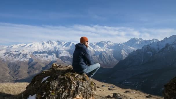 戴着太阳镜、戴着帽子的留着胡子的旅行者的肖像，坐在一块岩石上，背景是群山 — 图库视频影像