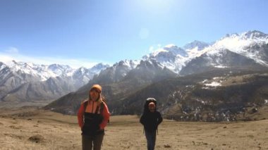 Şapka ve güneş gözlüğü sırt çantaları ile iki turist fotoğrafçılar karlı dağların arka planına karşı ellerinde kameralar ile sarı çim tepede yukarı gidin. yavaş hareket
