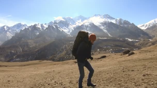 Due fotografi turisti con zaini in cappelli e occhiali da sole salgono sulla collina sull'erba gialla con le macchine fotografiche in mano sullo sfondo di montagne innevate. rallentatore — Video Stock