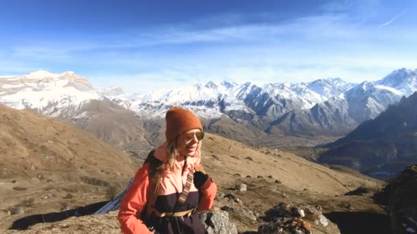 Feliz fotógrafa viajera en gafas de sol con una cámara y una mochila de pie cuesta arriba sobre un fondo de picos nevados en un día soleado — Vídeo de stock