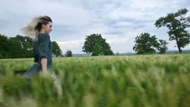 Fiatal boldog szőke lány fut egy zöld búza területén este ellen a háttérben az eső égen. Oldalnézet — Stock videók