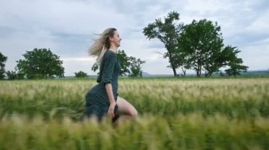 Genç mutlu sarışın kız yağmur gökyüzünün arka plan karşı akşam yeşil bir buğday tarlasında çalışır. Yan görünüm