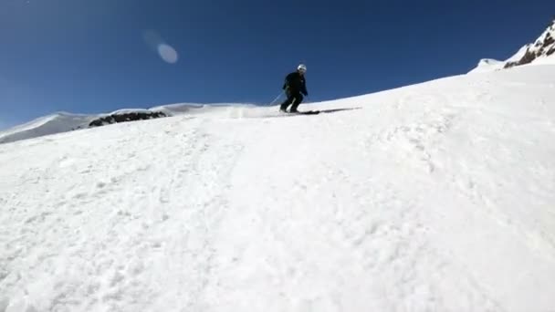 在阳光明媚的日子里，一名身着黑色装备、头戴白色头盔、带滑雪杆的广角男性滑雪者在雪坡上骑行。冬季滑雪运动的概念 — 图库视频影像