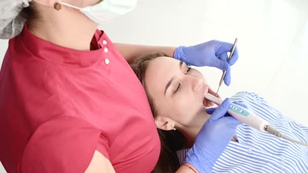 En professionell tandläkare kvinna i glasögon och overaller undersöker munhålan av en ung flicka i tandläkarstolen med hjälp av en intraoral stamotologisk videokamera med LED-belysning — Stockvideo