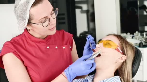 Mooi blond meisje in beschermende gele bril op de stamotoloog onderzocht haar open mond. Vrouwelijke tandarts onderzoekt de mondholte van een jonge patiënt met behulp van een tandarts-instrument — Stockvideo