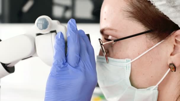 Close-up Uma mulher de óculos com um dentista profissional que trabalha com um microscópio estomatológico profissional em seu escritório. Conceito de profissão de estamotologista — Vídeo de Stock